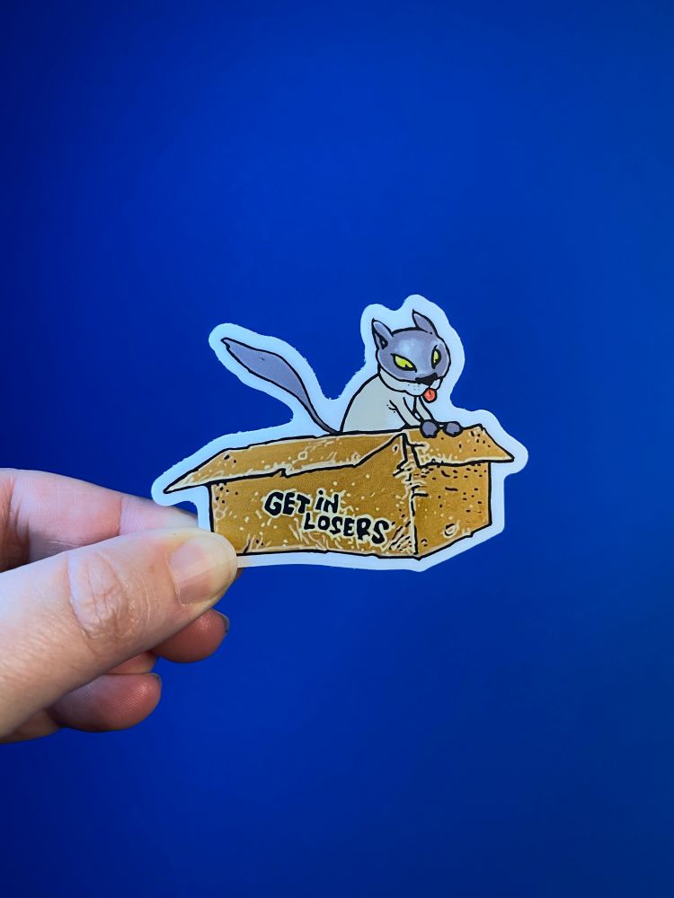 A hand holds a 3” vinyl sticker of a cat piloting a cardboard boxes labeled “Get in losers.” Artwork by Justin Groom.