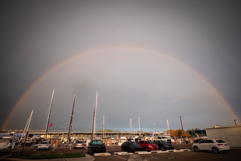 Full rainbow over boats in a marina and cars in a parking lot.