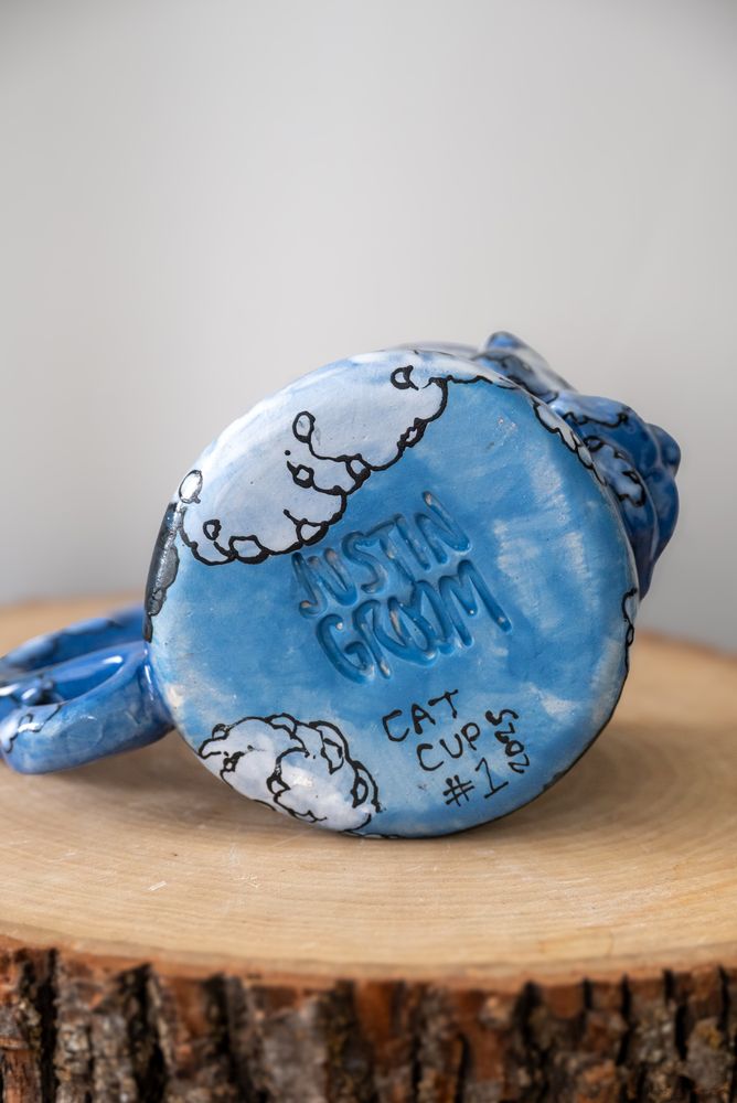 Handmade ceramic mug with raised cat face and tail-shaped handle, hand-painted in a cloud motif. This image shows the mug’s bottom, with artist’s signature. 