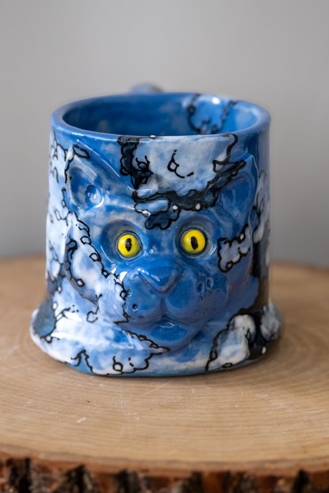Handmade ceramic mug with raised cat face and tail-shaped handle, hand-painted in a cloud motif.