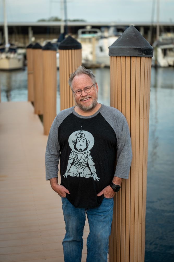 A smiling male artist leans against a post on a boat slip, wearing a t-shirt featuring his “Space Monkey” design. Water and boats in the background. 