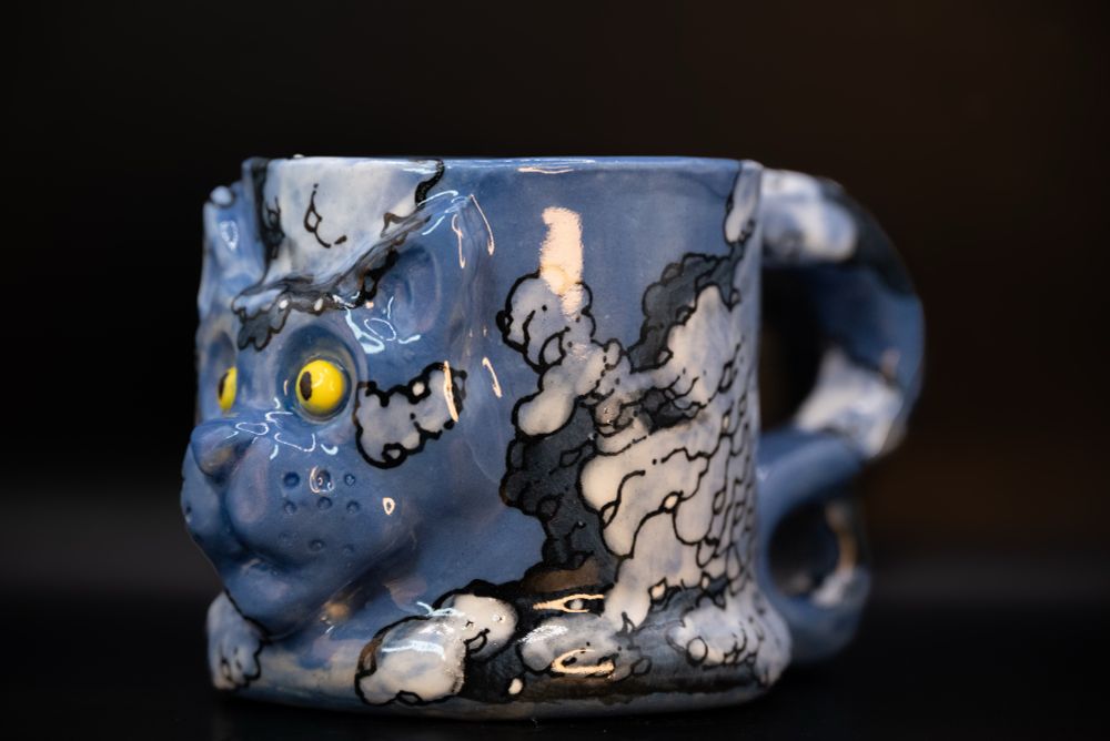 Handmade ceramic mug with raised cat face and tail-shaped handle, hand-painted in a cloud motif.