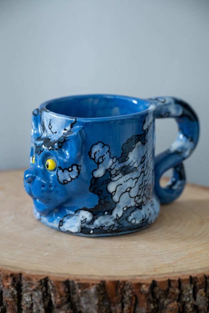 Handmade ceramic mug with raised cat face and tail-shaped handle, hand-painted in a cloud motif.