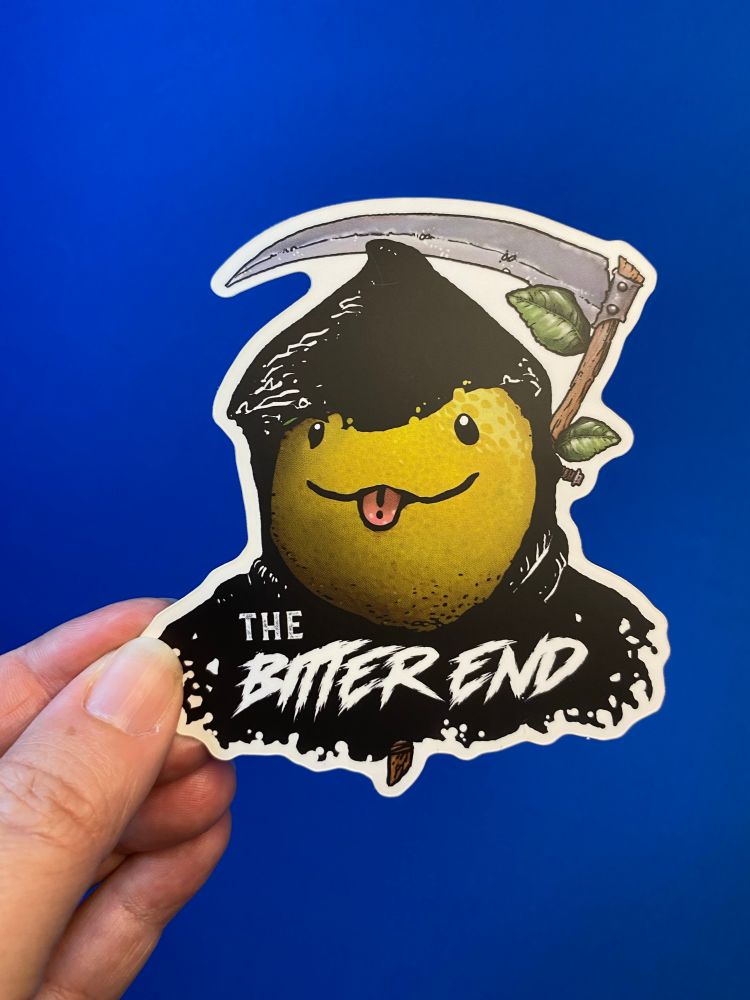 A hand holds up a sticker of a kawaii lemon grim reaper against a blue background.