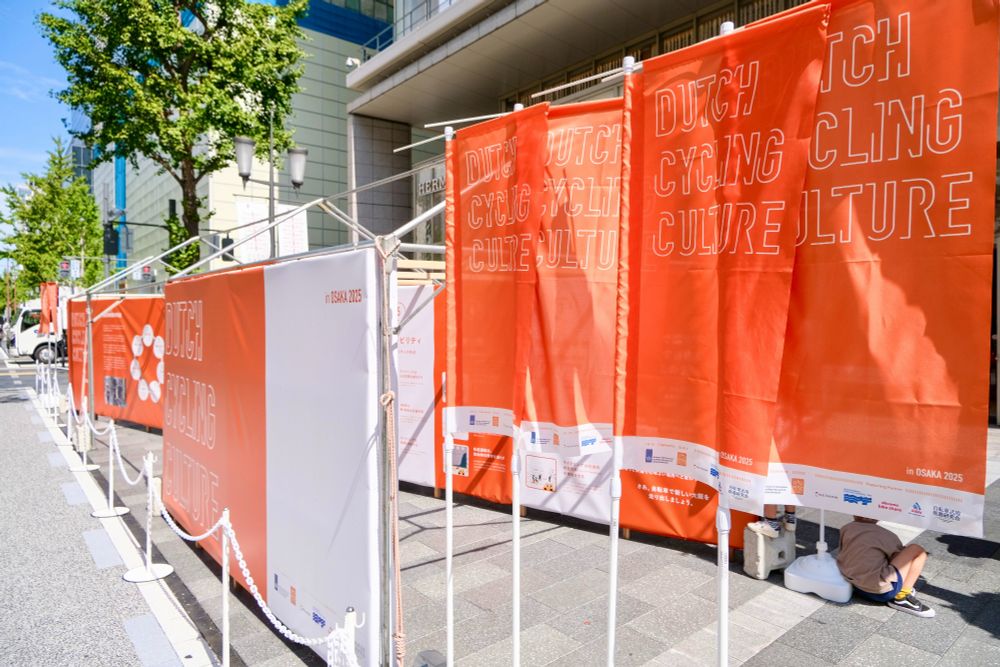 Photos of an orange and white exhibition consisting of photos and Japanese characters, on the theme “Dutch Cycling Culture”, set up along a busy foot and cycle path in the centre of Osaka.