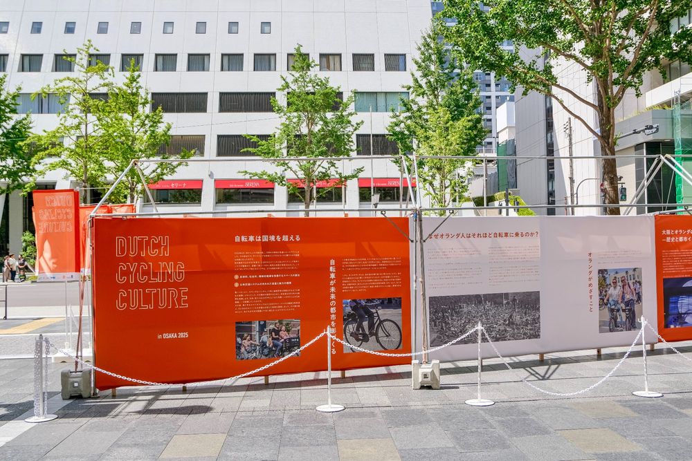 Photos of an orange and white exhibition consisting of photos and Japanese characters, on the theme “Dutch Cycling Culture”, set up along a busy foot and cycle path in the centre of Osaka.
