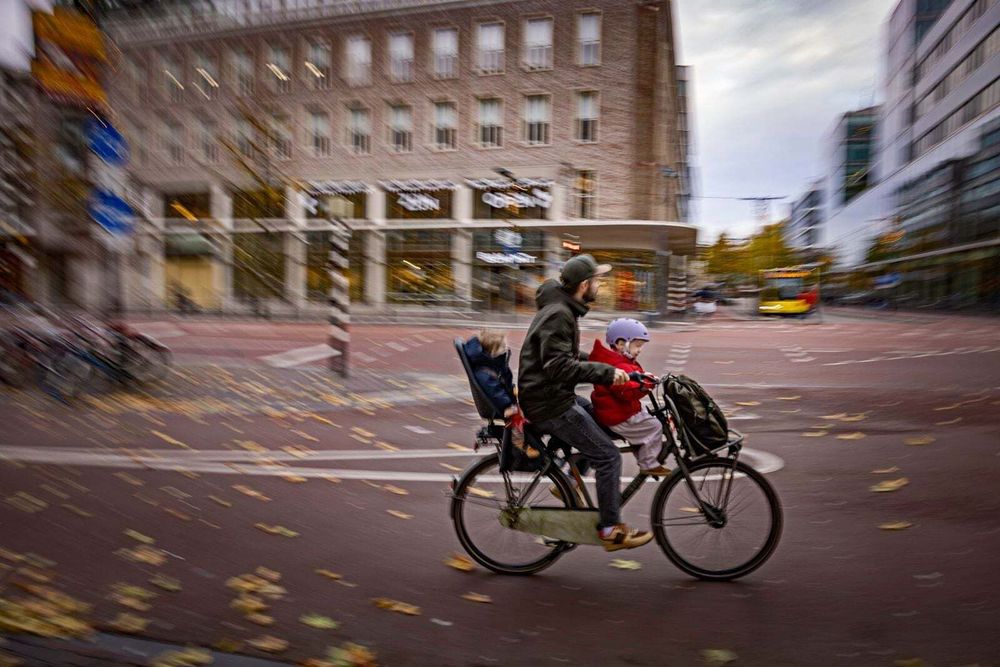 Man cycling by with blurred background and two children on front and back of bike

