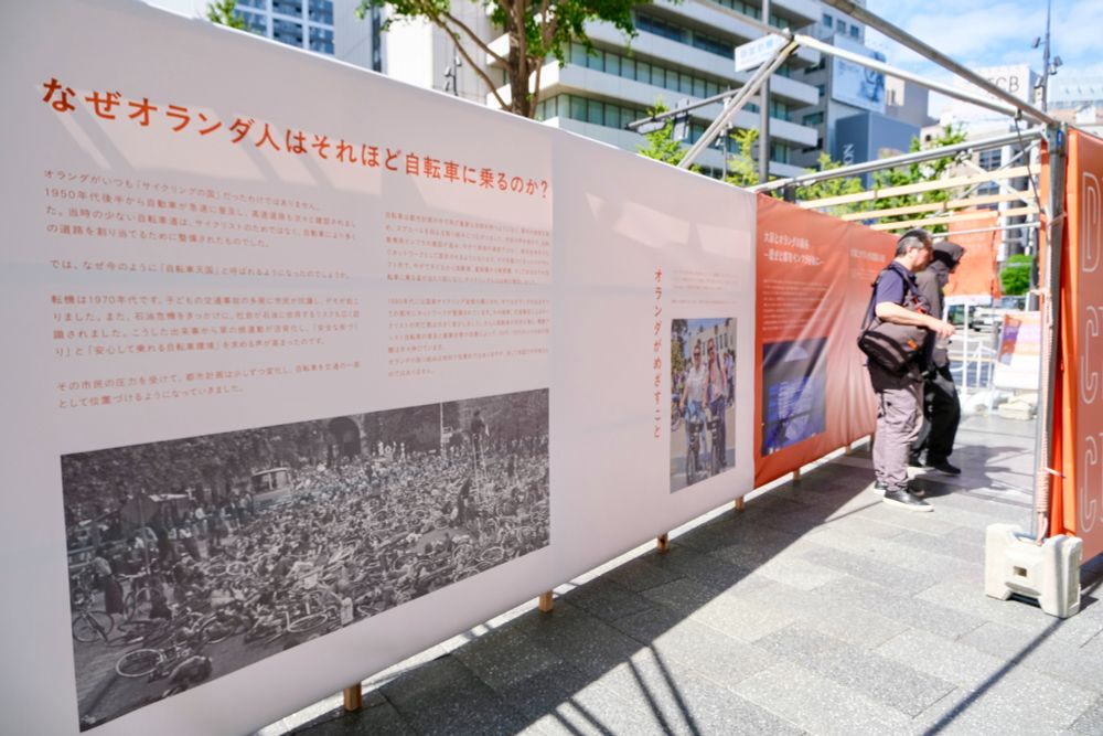 Photos of an orange and white exhibition consisting of photos and Japanese characters, on the theme “Dutch Cycling Culture”, set up along a busy foot and cycle path in the centre of Osaka.
