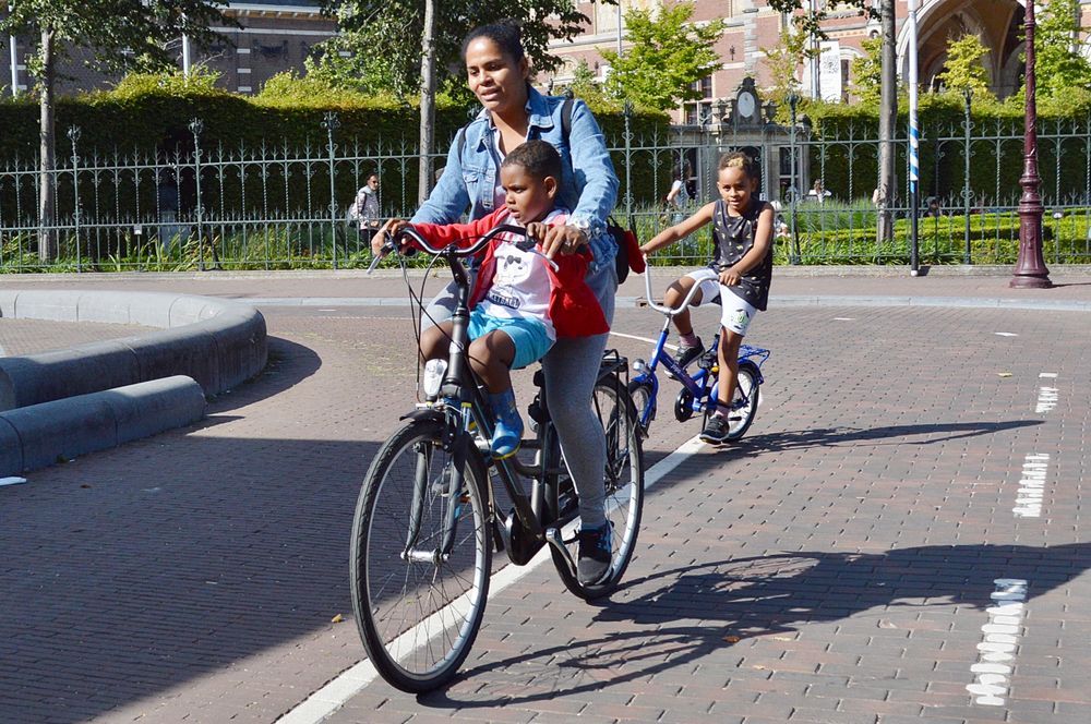 Mother riding her bike with children, one sitting on the frame, one riding behind.