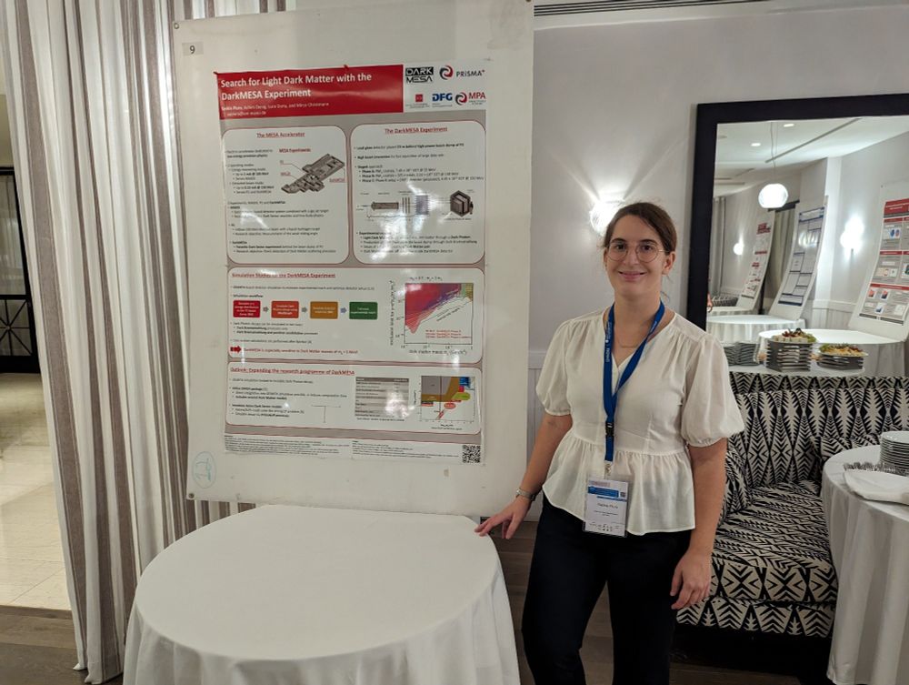 A young woman with brown hair, wearing a white blouse and dark blue dress pants, is standing next to a table. On the table a poster wall is placed, to which her poster is attached. The title of the poster says "Search for Light Dark Matter with the DarkMESA Experiment" and shows the current status of her research, which is about the simulation of the experimental reach of DarkMESA for different Dark Matter models.