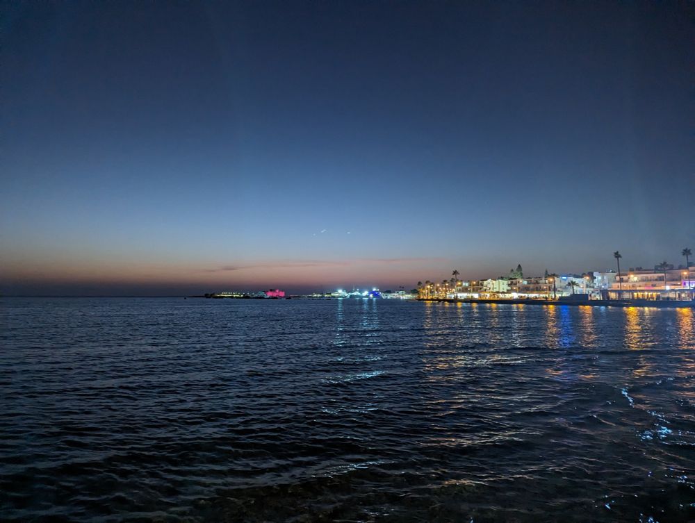 The photo shows the pier of Paphos during sunset. The lights of the pier are reflected in the Mediterranean sea.