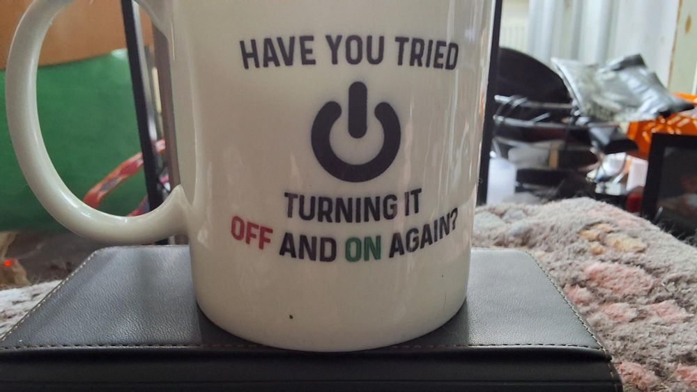 De afbeelding toont een witte mok met de tekst "HAVE YOU TRIED TURNING IT OFF AND ON AGAIN" (deze laatste zin in rood onder een aan/uit-symbool).