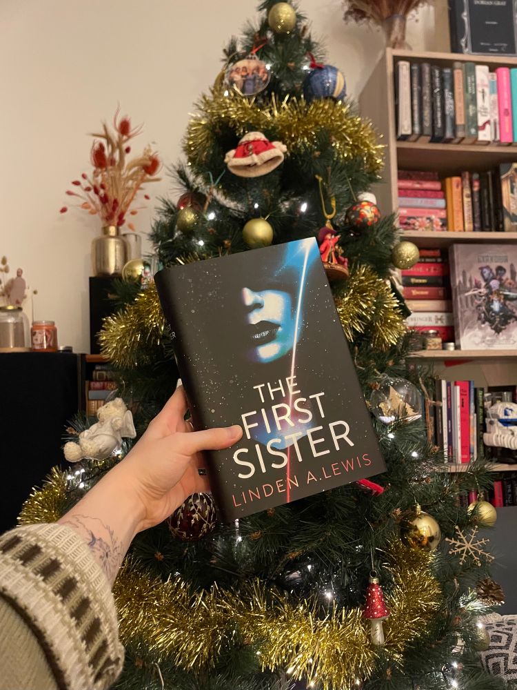photo of my hand holding the book the first sister by linden a. lewis in front of a christmas tree