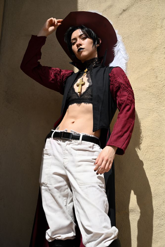 Genderbend cosplay photo of self as Dracule Mihawk. Photo taken from lower angle, 3/4 of the body looking down at camera. Outfit is same burgundy sleeve/black coat + hat combo as original, with the addition of a black lace bra top with kogatana connected via jump ring.