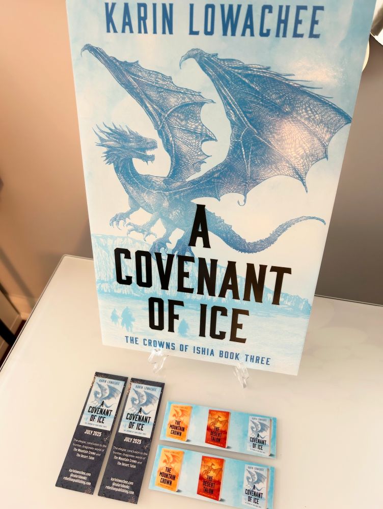 placard of book cover “a covenant of ice” by karin lowachee, blue with a dragon in flight. below it a spread of bookmarks, the book is out july 29