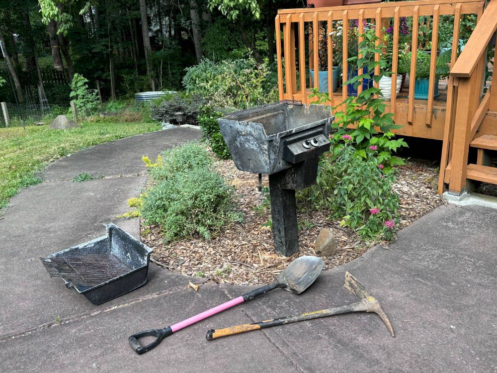 photo of an old crusty grill in a plant bed next to a shovel and pickaxe.