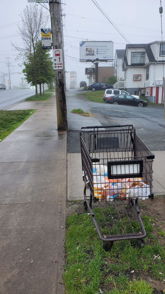 It's a sidewalk with a power pole. On the power pole is a bus stop sign. A few meters from the pole is a shipping cart full of garbage. In the background are a couple buildings with cars parked out front.