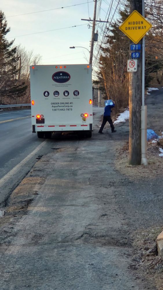 A large water delivery truck pulls up onto a sidewalk to park, blocking 85% of it.