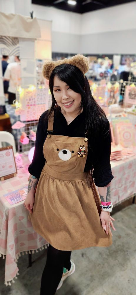 Merry, a long black haired Asian femme human in teddy bear overalls dress and a fuzzy teddy bear headband. Standing in front of their stall of art prints, stickers, keychains and furbies. The stall is lit up with star string lights.