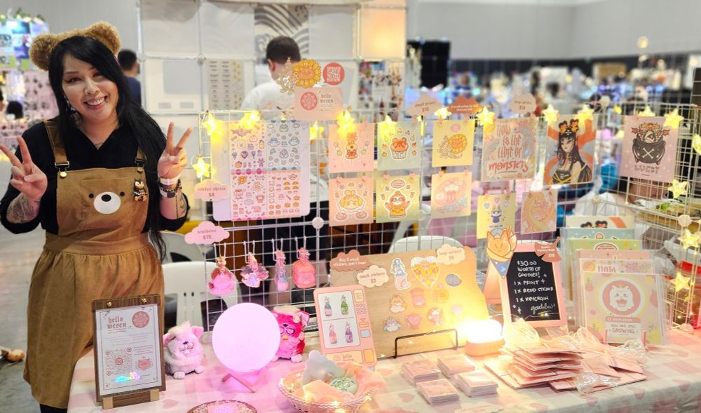 Merry, a long black haired Asian femme human in teddy bear overalls dress and a fuzzy teddy bear headband. Standing next to their stall of art prints, stickers, keychains and furbies. The stall is lit up with star string lights and a pink moon lamp.