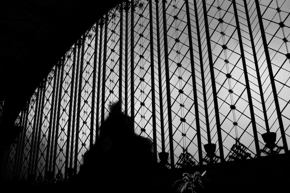 black and white image taken from inside the train station, looking at a huge semi-circular window. A structure of vertical lines and decorative trim make an interesting black pattern against the gray sky through the glass, and the silhouette of a building takes up part of the window as well.