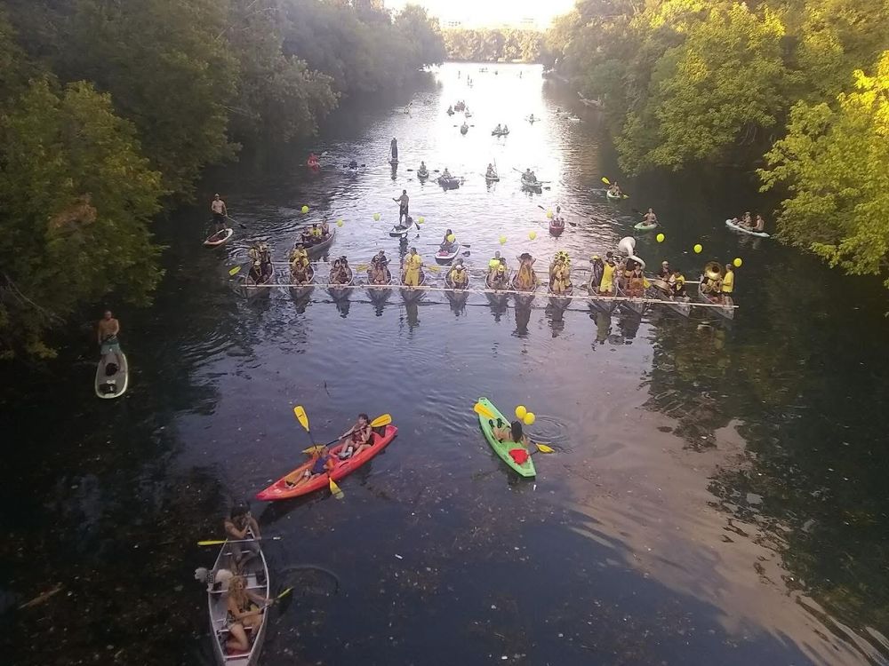 Picture of a marching band playing in 14 canoes strapped together on the water, so that they stretch all the way across the creek. The band is wearing black and yellow, and playing for a lot of people in boats and kayaks spread around the flotilla of canoes. 