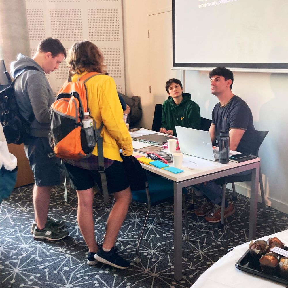A picture of students attending a technology exhibition. The picture displays two law students, one wearing a yellow hoodie and another wearing a grey hoodie talking to members of staff from Hive IT at their exhibition table. On the table is laptops, post-it notes and pens. 
