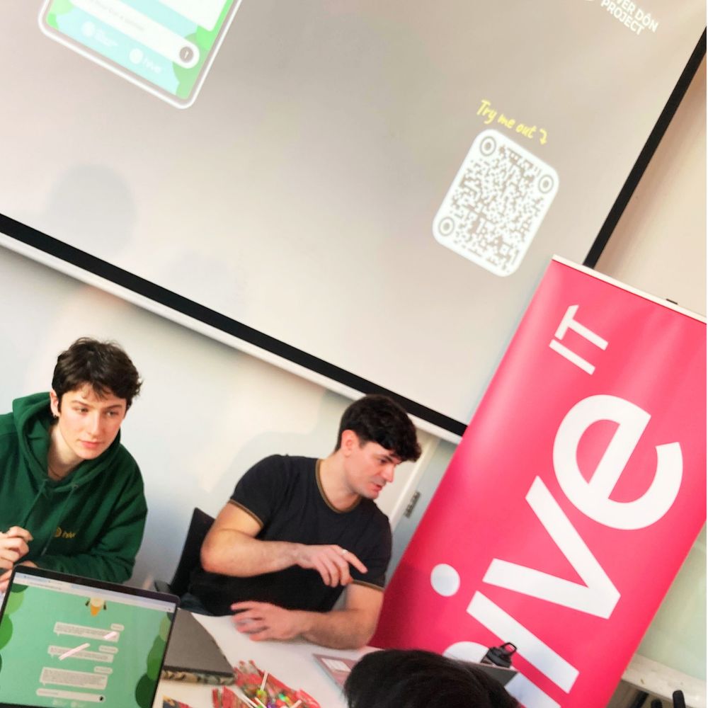 The image is a picture of Effy our user researcher wearing a green Hive IT hoodie, in deep conversation with a student from the university. Next to her is Alban wearing a black top, also in deep conversation with another student. There is also a pink advertisement sign next to Alban which says, 'Hive IT' and on the white wall behind them there is a projected image of a mobile app. 