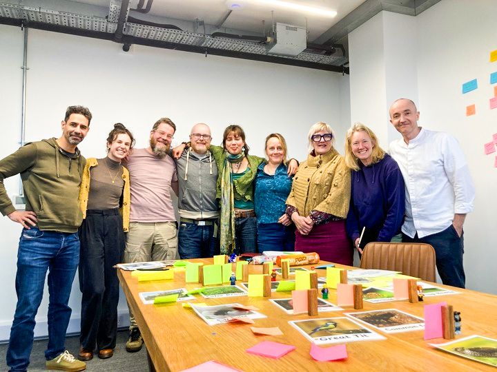 Group photo of Hive IT, Nature Nurture and A Noisy Silence stood together in a meeting room all smiling at the camera. On the table in front is lots of pictures and different coloured sticky notes to portray a miniature version of the ‘Web of Life Quest’ game so far.