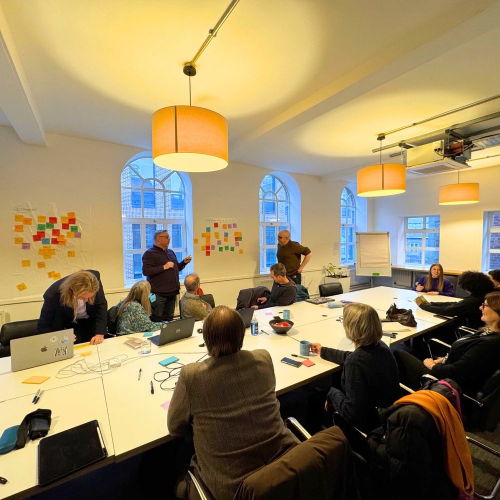 A group photo of people in a big meeting room taking part in a workshop facilitated by Hive IT. There is a big long table with laptops, markers, post-it notes and people sat on office chairs. On the walls of the room there is multi-coloured post-it notes placed on the wall as a workshop exercise. 