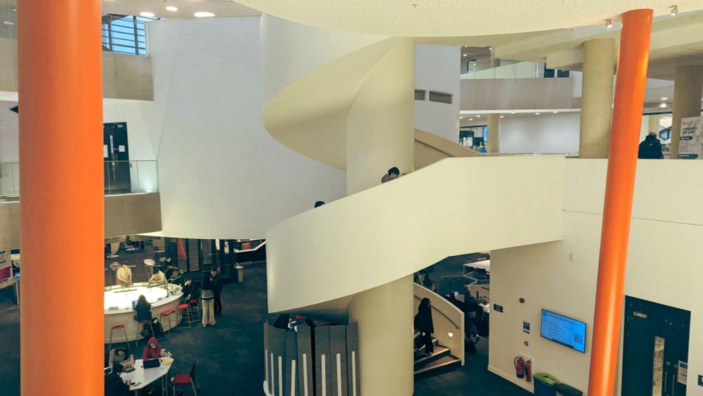 A picture of inside the Diamond building at the university of Sheffield. Students are studying on tables and there is a spiral staircase in the middle of the picture.