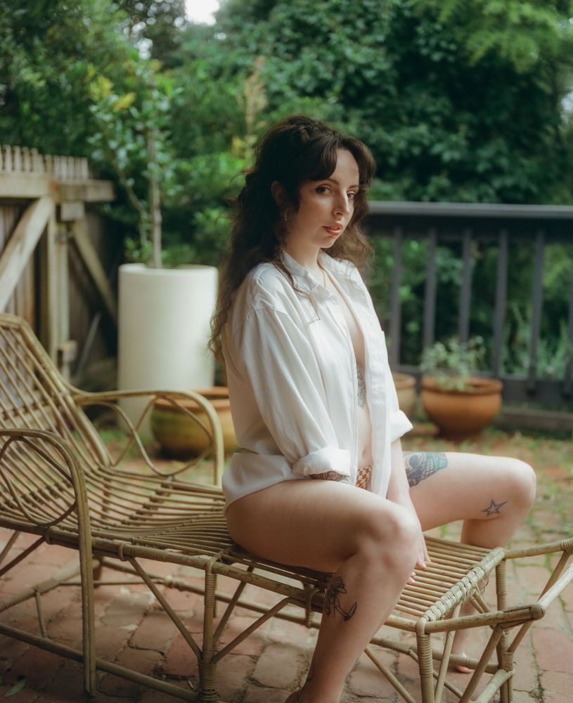 Hailey sits on a wicker bench in an outdoor garden setting, wearing an oversized white button-down shirt draped casually over her shoulders. Surrounded by lush greenery and potted plants, she gazes at the camera with a calm, confident expression. The scene has a soft, romantic ambiance with natural light filtering through the foliage.