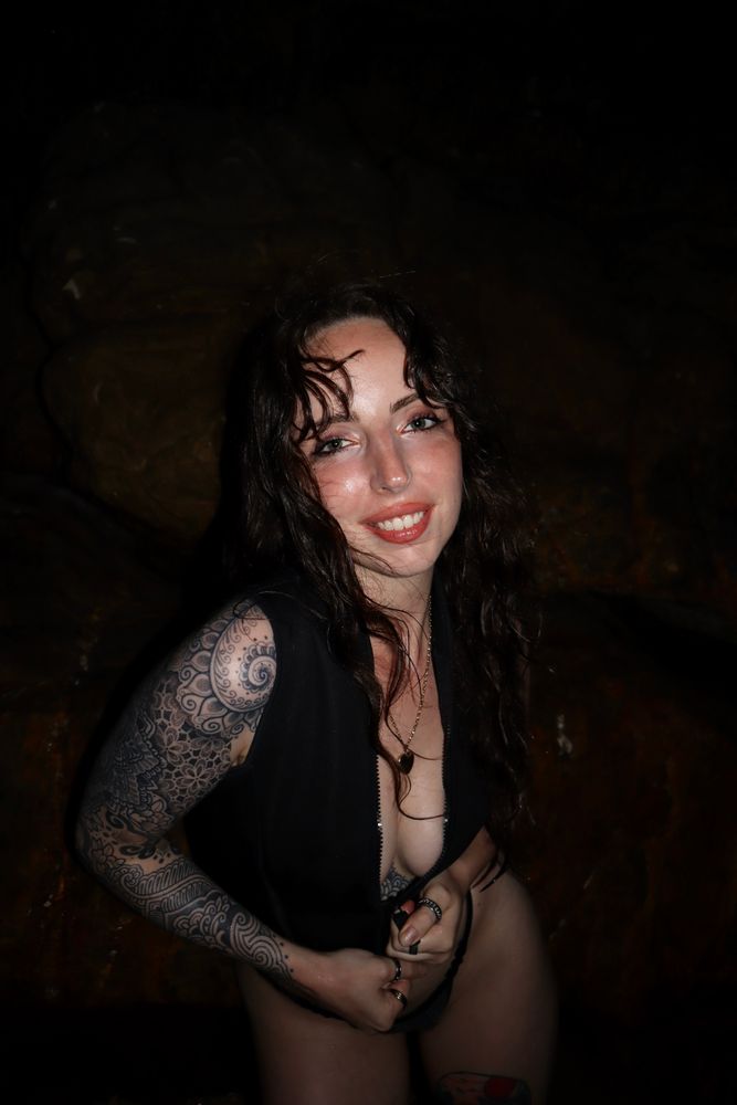 Hailey Hendrix stands at night with her back to a dark, rocky wall, her wet hair tousled and falling around her face. She’s wearing a black zip-up swimsuit, slightly unzipped to reveal the top of her chest tattoo. Her full sleeve tattoo on her right arm is also visible. She’s smiling warmly at the camera with both hands resting near her hips, fingers loosely interlaced. Her skin glistens under the camera flash, suggesting she’s recently been in the water. The mood is intimate, playful, and confident.