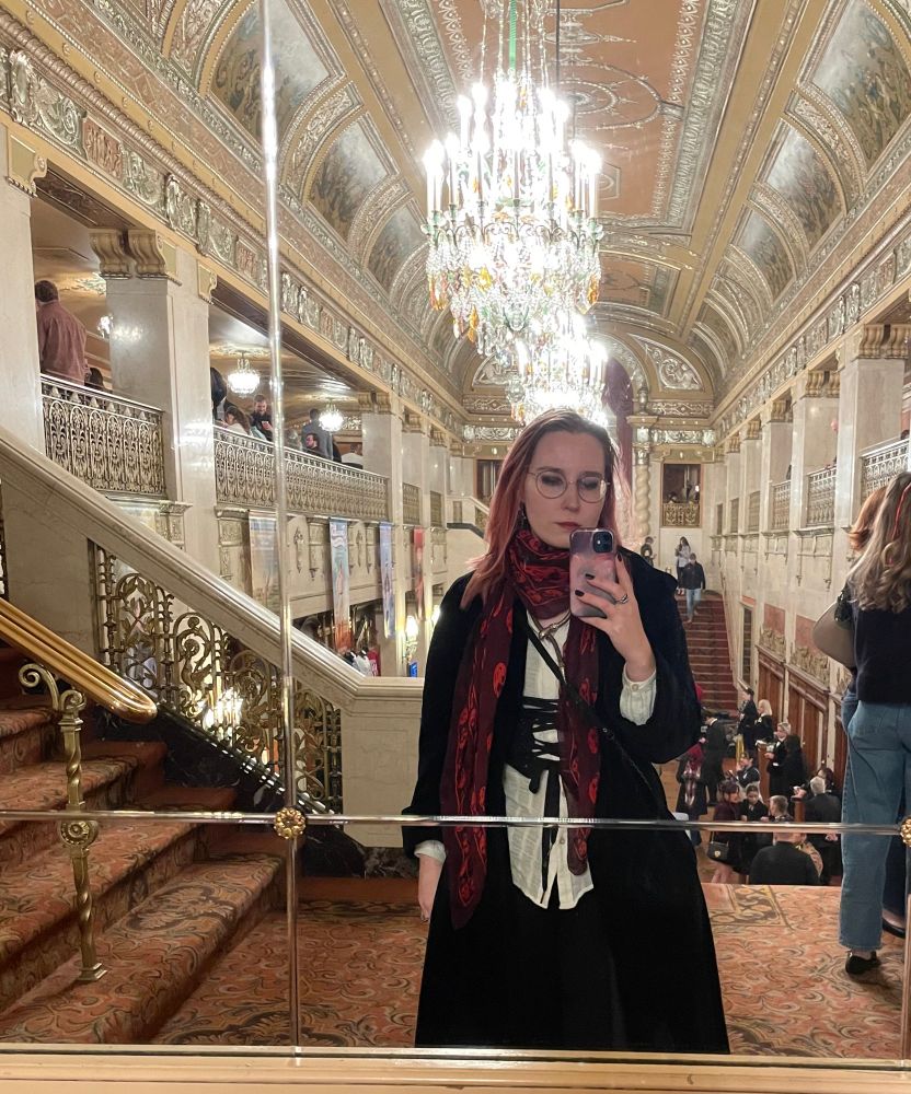 Self portrait in a mirror. Rows if chandeliers run the ceiling behind me. I’m wearing a black velvet coat, a red McQueen skull scarf, a white shirt, and a black corset-front bustier/bodice thing. 