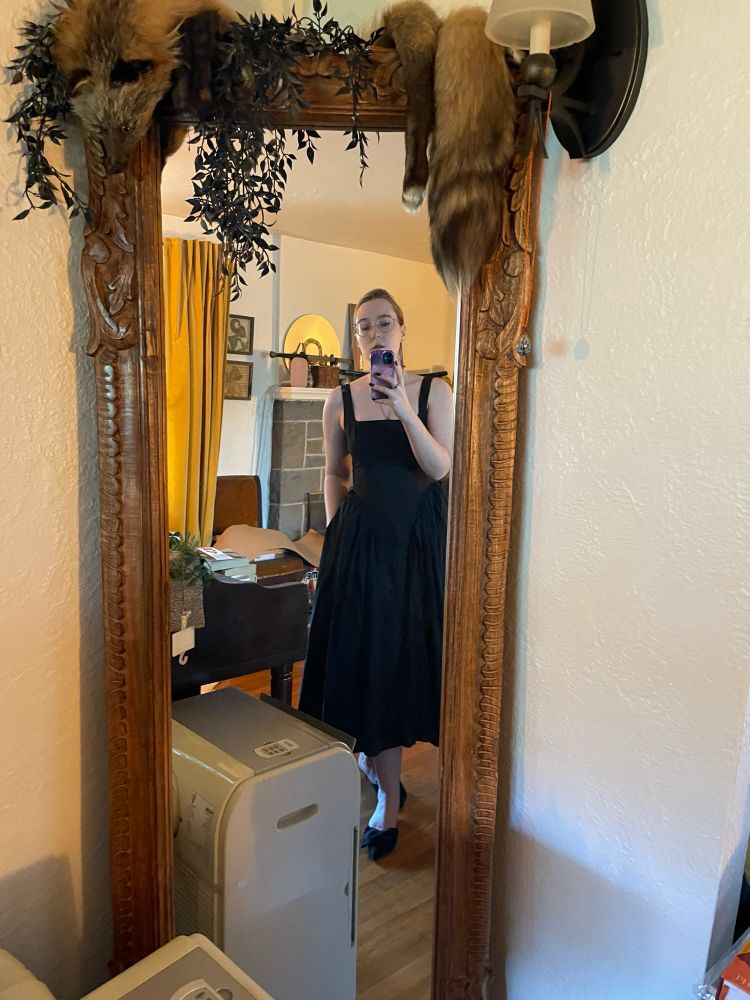 Selfie of a white woman in a midi black dress, with pointy black shoes. The floor length mirror has a craved wood frame with a vintage fox stole draped over it. A floor air conditioning unit is taking up half the bottom of the mirror, which is very annoying
