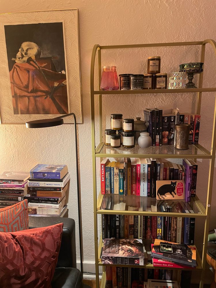 A gold and glass open bookshelf standing next to a couch, behind which more books are stacked. The top two shelves are mostly jar candles. An oil painting of a skull hangs on the wall next to it. 