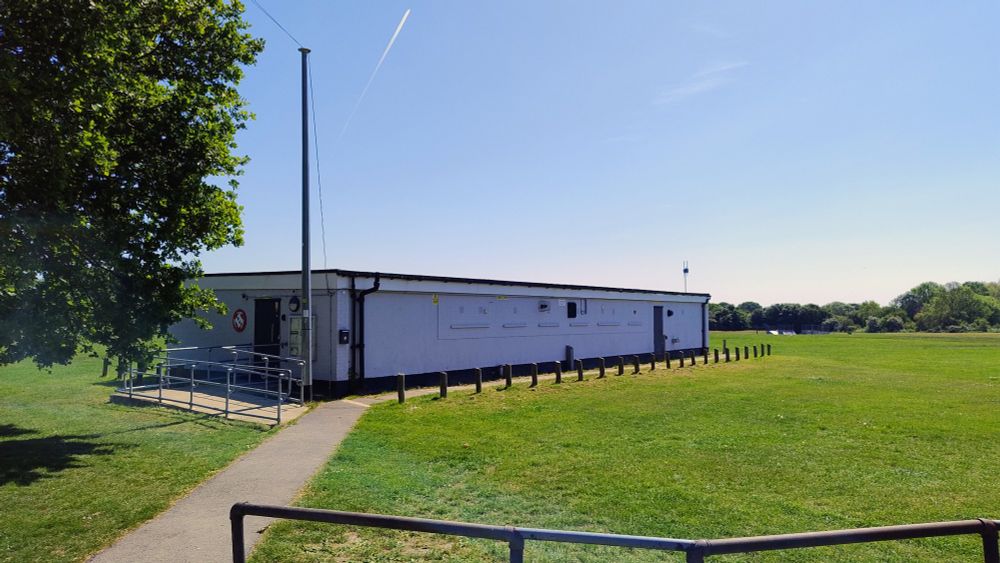 A picture of the low square Hackspace building in bright sunshine. The grass on the field looks recently mowed.