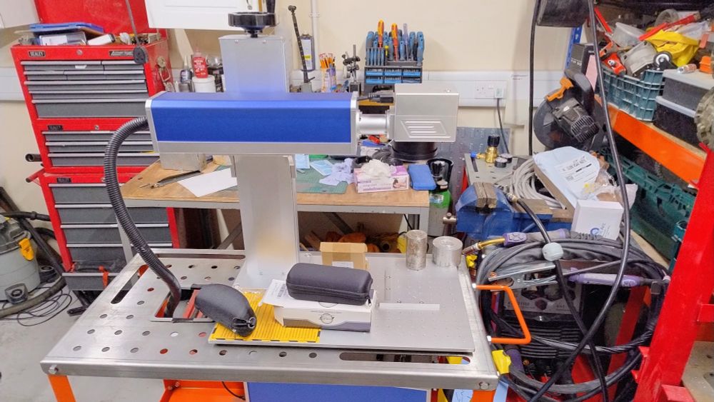 The head unit of our MOPA laser engraver sat on a wiring table. A square hole has been cut in the top to allow the big cable bundle that includes the fibre can pass down to the laser module that sits on the shelf below. The head unit is a chunky square body raised above a large aluminium need used to hold the workpiece. This can be wound up and down to get the focus right with a handwheel. It sort of looks like a prop from Star Trek TOS.