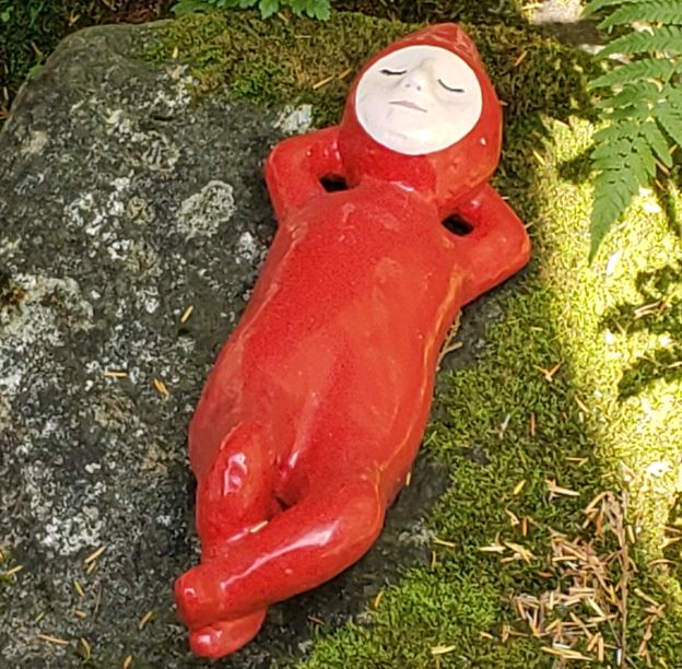 my profile picture, which is a photo of a little red ceramic guy with a pointy head and a circular white face, laid back and relaxing on a shaded mossy rock. 