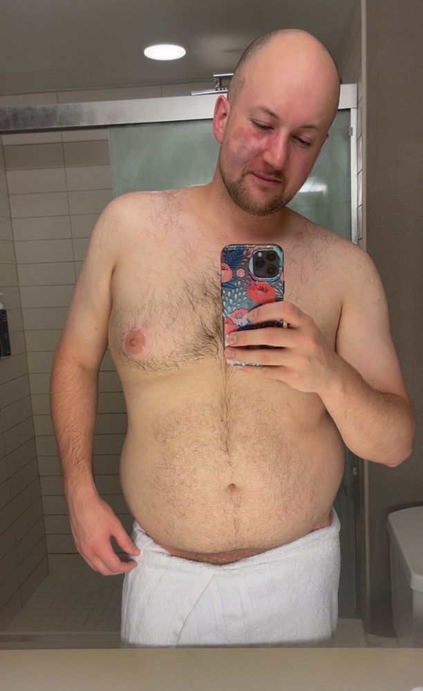 A man with a towel around his waist takes a mirror selfie 