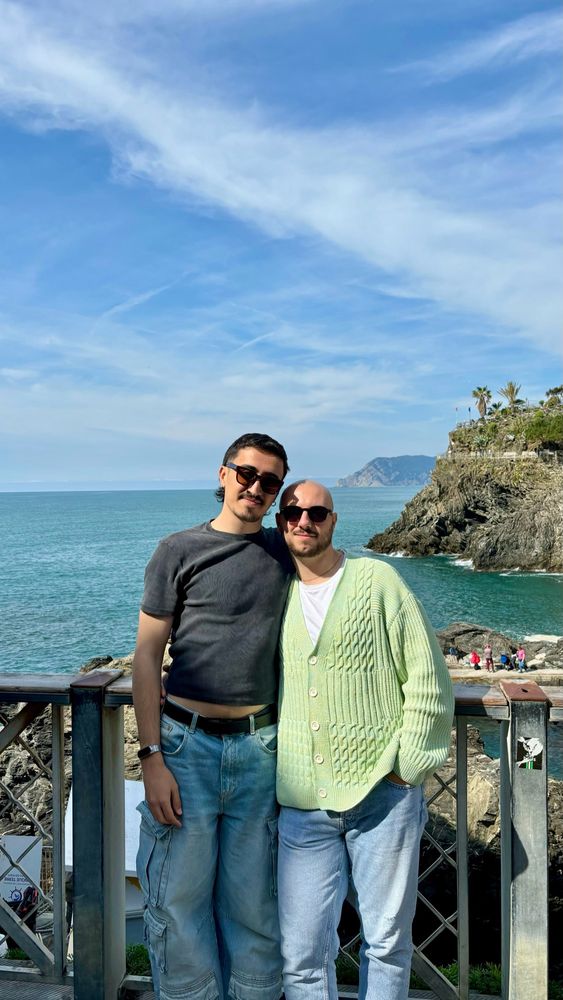 Ale y yo en Riomaggiore, Italia. Posamos frente a la cámara apoyados en la barandilla de un balcón. A nuestras espaldas está el mar y grandes piedras que conforman la cala alrededor de la cual se asienta la ciudad.