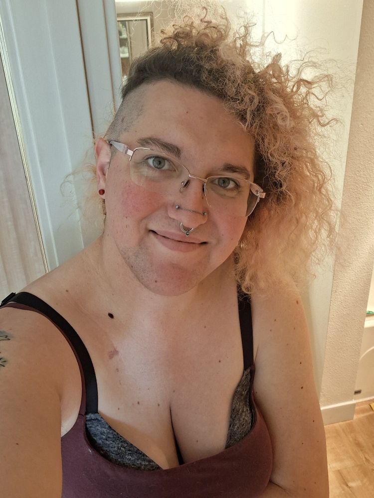 Trans girl with curly dyed blonde hair, pjnk glasses, nose piercings and her cleavage all the way out in a red tank top giving a warm smile.