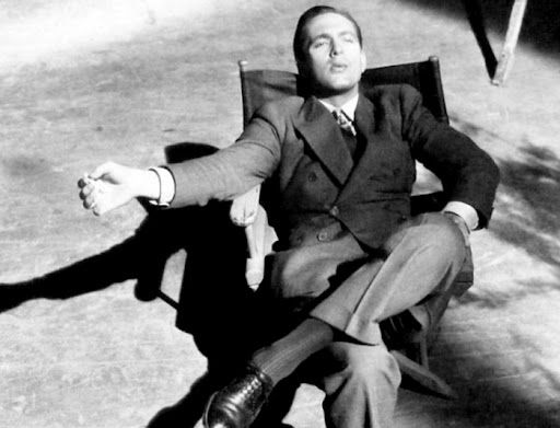 Fredric March on the set of Design for Living sitting cross-legged in a director’s chair, head back blowing smoke from a cigarette. He looks fine as hell