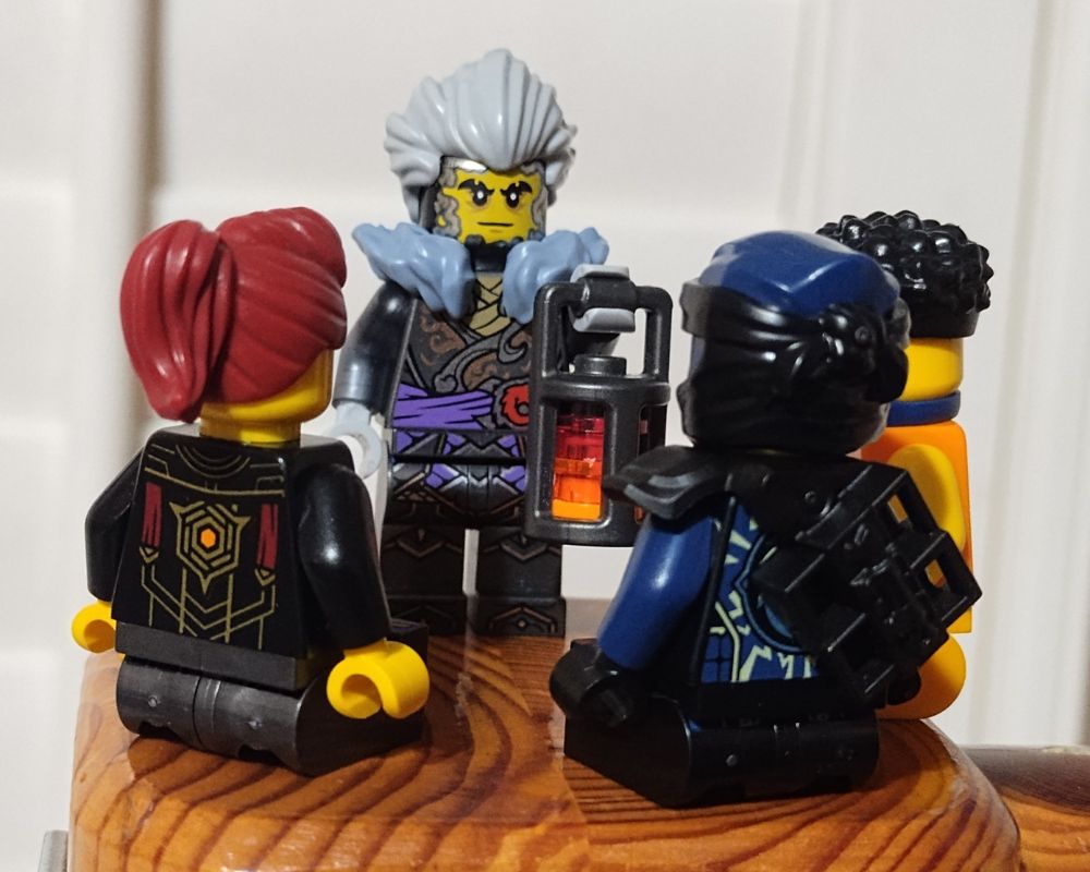 Jordana, Jay, and Arin are seated in an arc in front of Cinder, who is standing and speaking while holding a lantern.