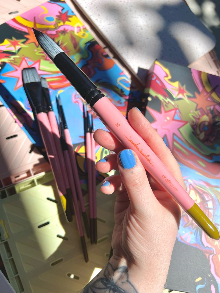 Pink paint brushes with black metal and chartreuse tip. The brush hairs are a grey to black gradient. The packaging is in the background.
