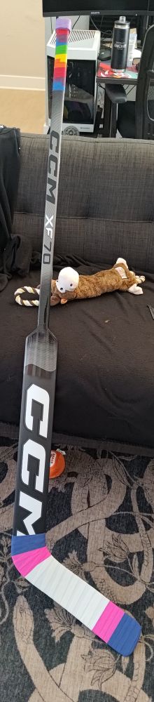 A CCM XF70 goalie stick for ice hockey. It's primarily black with grey gradients. It has rainbow tape near the top of it going up to a purple tape knob, and blue, pink and white tape in the pattern of the trans pride flag wrapping the blade from toe to heel.