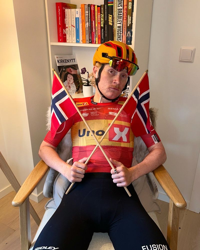 Magnus Kulset sat down in a chair, wearing his cycling kit, while holding two Norwegian flags. The two small flags are crossed over each other and he is pulling a slight pout. 
