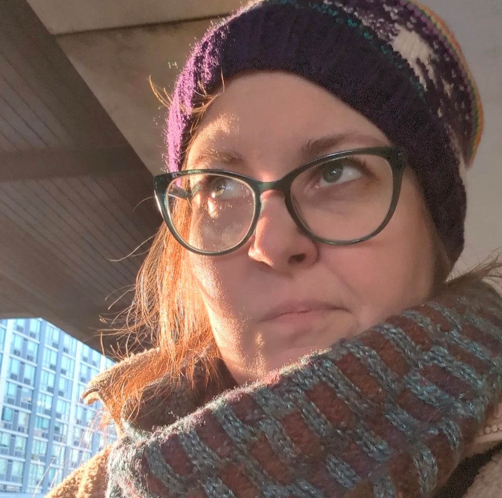 Grumpy Kates it outside on a train platform wearing snazzy glasses and hand knits, including a purple hat with unicorns and a multicolored cowl around her neck.
