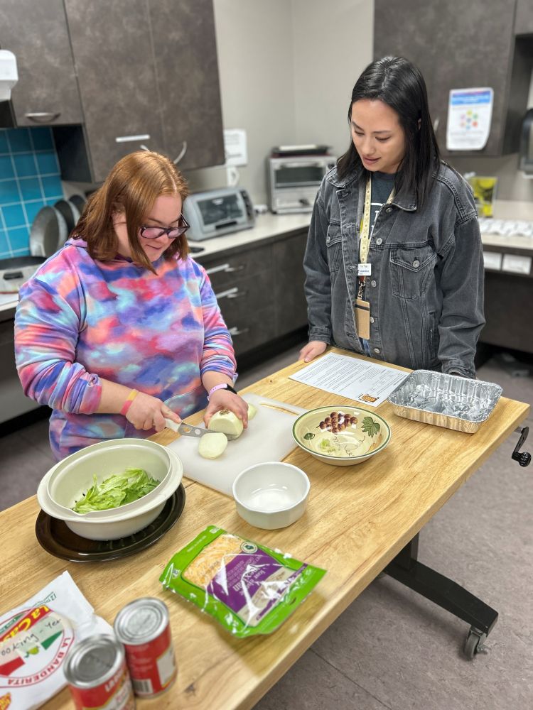 A young woman with a purple sweater slices onions on a cutting board. Another young woman is standing beside her watching.