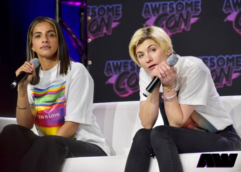 Mandip Gill and Jodie Whittaker on the stage of a convention, both holding microphones. Mandip is wearing a shirt reading "this is love"; Jodie has much shorter blonde hair than usual and is wearing many bracelets.